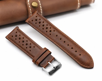 Könnte beinhalten: Ein braunes Lederuhrenarmband mit perforiertem Design und zahlreichen kleinen Löchern auf der Oberfläche. Das Armband verfügt über eine silberfarbene Metallschnalle und zwei passende Lederschlaufen sowie eine glatte Oberfläche mit sichtbaren Nähten.