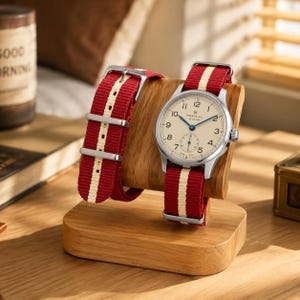 May include: A wristwatch with a cream-colored dial, blue hands, and a red and white striped nylon strap, displayed on a curved wooden stand. The watch face features the text Seagull and 19 Zuan, with Arabic numerals and a small sub-dial.