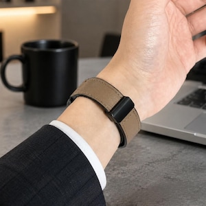 Könnte beinhalten: Ein taupefarbenes Leder-Uhrenarmband mit glatter, matter Oberfläche und sichtbaren, farblich abgestimmten Nähten an den Kanten. Das Armband wird mit einer rechteckigen schwarzen Metallschließe befestigt und bietet ein schlichtes Design.