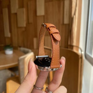 Op de afbeelding: Een smartwatch met een beige leren band met zichtbare stiksels en een zilverkleurige metalen gesp. De horlogekast is rond met een zwart achterpaneel, voorzien van een metalen afwerking en een slank, verstelbaar bandontwerp voor een goede pasvorm.