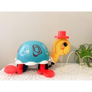 May include: A retro toy turtle with a blue shell and white and maroon details. The turtle has red feet, a yellow head with a red hat, and a black and white striped collar. A classic children's toy.