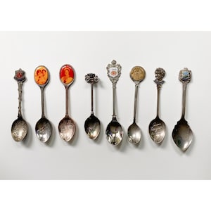 May include: A collection of eight silver souvenir spoons with various designs. The spoons feature images of a royal couple, a crown, a city skyline, and a portrait of a man.