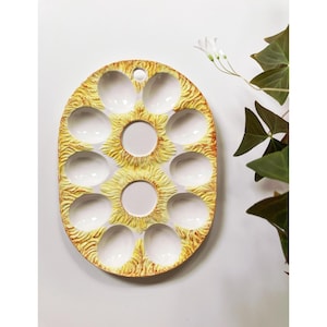 May include: A white ceramic egg holder with a yellow and orange design. The holder has 12 individual compartments for eggs.