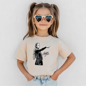 May include: A beige short-sleeved t-shirt featuring a black graphic print of a volleyball player in a jersey with the number 8, pointing to the side, accompanied by a signature. The shirt has a crew neckline and a standard fit.