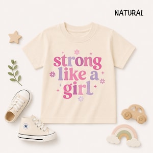 May include: A natural-colored short-sleeve t-shirt featuring the text "strong like a girl" in a pink and purple gradient font, accented with small star and flower graphics. The shirt is displayed alongside a pair of cream-colored sneakers.