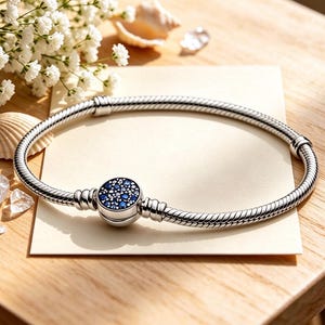May include: A silver snake chain bracelet featuring a circular clasp adorned with a cluster of small, sparkling blue and clear stones. The bracelet is positioned on a rectangular card, showcasing a polished metallic finish and a secure locking mechanism.