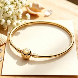 Puede incluir: Una pulsera rígida de tono dorado pulido con una banda circular lisa y un cierre esférico central grabado con el texto PANDORA. La pulsera presenta un diseño sencillo con dos pequeños detalles acanalados a los lados del cierre central.