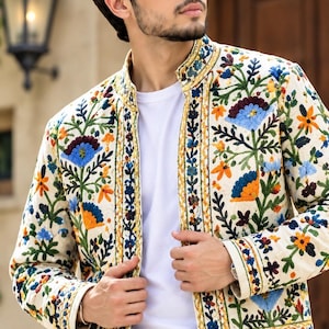 May include: An off-white jacket featuring a vibrant, multi-colored floral embroidery pattern in shades of blue, orange, and green. The garment has a mandarin collar, long sleeves, and a straight hem, worn over a plain white t-shirt.