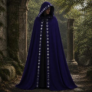Puede incluir: Una capa con capucha de color morado oscuro que llega hasta el suelo, con un patrón de fases lunares bordado en plata a lo largo de los bordes delanteros y el borde de la capucha. La prenda se lleva sobre una base negra lisa y fluida.