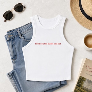 May include: A white sleeveless crop top featuring the red text Pretty on the inside and out printed on the front. The garment is displayed alongside blue denim jeans, black sunglasses, a straw hat, and a book with a partial title visible.