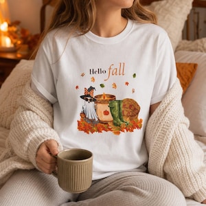 Könnte beinhalten: Ein weißes Kurzarm-T-Shirt mit einem Grafikdruck und dem Text Hello fall. Das Motiv zeigt ein Gespenst mit Hut und Sonnenbrille, eine Kerze, grüne Gummistiefel und einen Kürbis, umgeben von verstreuten herbstlichen Blättern.