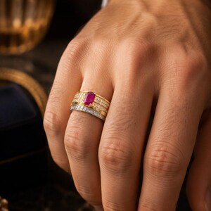 May include: A multi-band ring set featuring a central oval-cut vibrant red ruby gemstone flanked by two yellow gold bands encrusted with small clear diamonds, paired with a separate silver-toned band featuring a pavé setting of clear diamonds.
