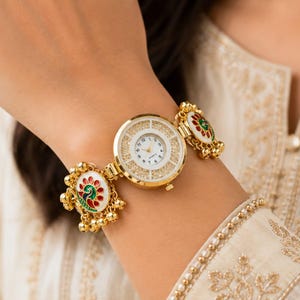 Könnte beinhalten: Eine goldfarbene Analogarmbanduhr mit rundem weißem Zifferblatt und schwarzen Ziffern. Das Armband ist mit zwei runden, weiß emaillierten Anhängern verziert, die ein buntes Pfauenmotiv und kleine baumelnde goldfarbene Metallglöckchen zeigen.