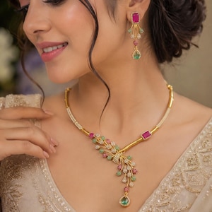 May include: A gold-toned necklace and matching drop earring set featuring small white, pink, and green stones arranged in a floral-inspired cluster. The necklace includes a prominent side pendant with a teardrop-shaped green stone at the base.