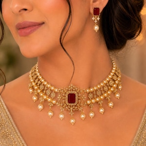 May include: A gold-toned choker necklace featuring a central rectangular deep red gemstone surrounded by intricate filigree metalwork, accented with multiple rows of cream-coloured pearls and dangling pearl drops, paired with matching drop earrings.