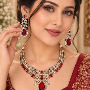 May include: A jewelry set consisting of a necklace and matching drop earrings. The necklace features a multi-layered design with gold-tone metal, clear crystals, and deep red teardrop-shaped stones. The earrings mirror the necklace's design.