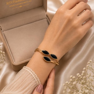 Könnte beinhalten: Ein goldfarbenes, offenes Armband mit zwei schwarzen Einlagen in Markisenform an den Enden. Das Armband wird neben einer beigen Schmuckschatulle präsentiert, auf deren Innendeckel der Text RSVFashionJewels in Gold aufgedruckt ist.