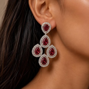 Puede incluir: Un pendiente tipo candelabro con cuatro piedras de color rojo intenso en forma de pera dispuestas en racimo. Cada piedra está rodeada por un halo de pequeñas piedras claras y brillantes en un marco de metal plateado.