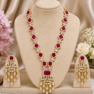 Puede incluir: Un collar y un par de pendientes a juego en tono dorado con piedras rectangulares de color rosa fucsia rodeadas de pequeños cristales transparentes. El collar tiene un colgante central con diseño de flecos y los pendientes siguen el mismo estilo.