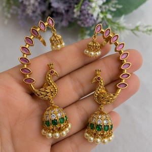 Peut inclure: Une paire de boucles d'oreilles dorées en forme de paon avec des pierres roses taille marquise le long de l'arc. Chaque pièce comprend un pendentif jhumka en forme de cloche orné de cristaux blancs, de pierres vertes et de petites perles.