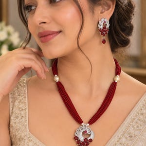May include: A multi-strand necklace featuring deep red beads, accented with gold-toned spacers and a central silver-toned pendant set with red teardrop stones and clear crystals, paired with matching drop earrings in a similar design.