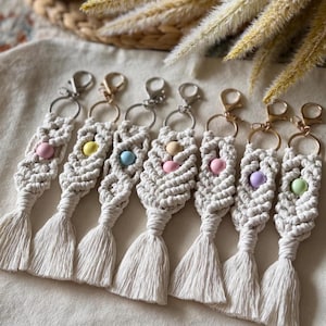 May include: Seven macrame keychains featuring off-white knotted cord, each with a single round pastel-coloured bead in shades of pink, yellow, blue, peach, lavender, and mint, finished with a metal lobster clasp and a short fringe tassel.