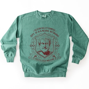 May include: A teal crewneck sweatshirt featuring a dark red graphic of a philosopher with a circuit board pattern on half of the face, surrounded by binary code, the text SILICON VALLEY CAMPUS DEPT. OF AI & NEURAL NETWORKS, and COGITO ERGO MACHINE.