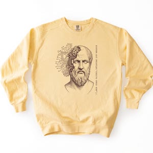 May include: A pale yellow crewneck sweatshirt featuring a dark brown graphic of a classical bust with a mechanical brain composed of gears and circuit board patterns, accompanied by the text: SYSTEM.OUT.PRINTLN("COGITO ERGO SUM");