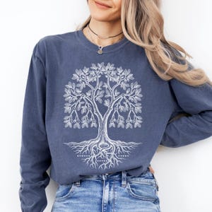 May include: A long-sleeve crewneck sweatshirt in a faded denim blue color. The front features a white graphic print of a stylized oak tree with intricate roots and branches. Small circles containing numbers are integrated into the tree design.