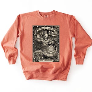 May include: A coral-colored crewneck sweatshirt featuring a black and white graphic print of a hooded figure working at a desk with alchemical symbols, a glowing geometric shape, and the text "TRANSMUTATIO DIGITALIS" and "THE ALCHEMIST".
