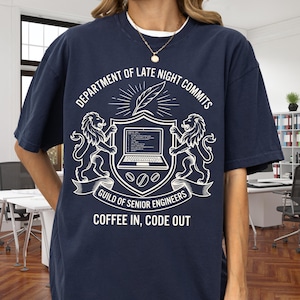 May include: A navy blue t-shirt featuring a white graphic of a crest with two lions, a laptop, and a quill. The text reads: DEPARTMENT OF LATE NIGHT COMMITS, GUILD OF SENIOR ENGINEERS, and COFFEE IN, CODE OUT. The design includes coffee bean icons.