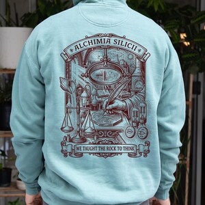 Op de afbeelding: Een lichtblauwe hoodie met een donkerrode afbeelding op de achterkant. De illustratie toont een alchemistisch laboratorium met een hand die op een printplaat schrijft, een balansweegschaal en vacuümbuizen. De tekst luidt ALCHIMIA SILICII en WE TAUGHT THE ROCK TO THINK.
