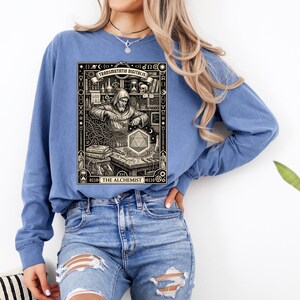 May include: A blue long-sleeve sweatshirt featuring a black and cream graphic print of a hooded figure working at a desk with alchemical symbols, a glowing geometric shape, and the text "TRANSMUTATIO DIGITALIS" and "THE ALCHEMIST" at the top and bottom.