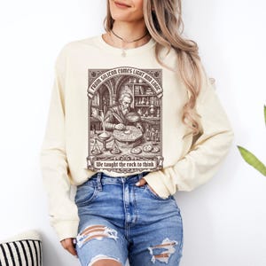 May include: A cream-colored long-sleeve sweatshirt featuring a dark brown graphic print of a scholar working at a desk with laboratory equipment. The text reads: FROM SILICON COMES LIGHT AND LOGIC and We taught the rock to think.