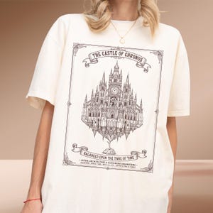 May include: An off-white short-sleeve t-shirt featuring a dark brown line art illustration of a castle with clockwork details. The text reads THE CASTLE OF CHRONOS and BALANCED UPON THE TWIG OF TIME, with additional architectural labels at the bottom.