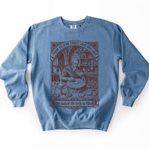 Puede incluir: Una sudadera azul de cuello redondo con una ilustración gráfica de color marrón oscuro de una persona barbuda trabajando en un escritorio con herramientas científicas. El texto dice "FROM SILICON COMES LIGHT AND LOGIC" y "We taught the rock to think".
