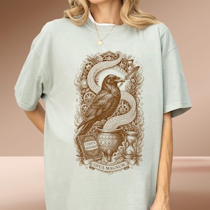 May include: A light sage green t-shirt featuring a brown graphic print of a raven perched on a cauldron. The design includes a quill, an hourglass, gears, and a book labeled CODEX BINARII, with a banner at the bottom reading OPUS MAGNUM.