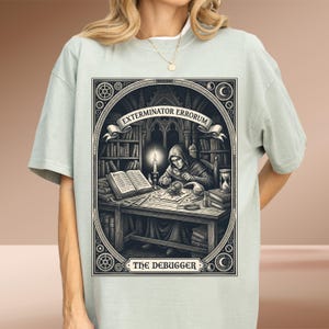 May include: A light sage green t-shirt featuring a graphic print of a hooded figure at a desk examining a beetle with a magnifying glass. The illustration includes books, a candle, and an hourglass, with the text EXTERMINATOR ERRORUM and THE DEBUGGER.