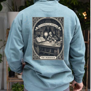 May include: A light blue hooded sweatshirt featuring a rectangular graphic on the back. The illustration depicts a hooded figure at a desk in a library, with the text EXTERMINATOR ERRORUM and THE DEBUGGER printed in a decorative frame.