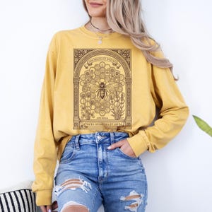 May include: A mustard yellow long-sleeve sweatshirt featuring a dark brown graphic print of a bee centered within a honeycomb pattern, framed by an ornate archway. The text reads APIS REGINA ET NATURA, APIS REGIA, and SACRA GEOMETRIA ET ARS.