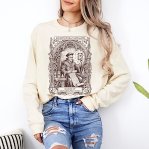 May include: A cream-coloured long-sleeve sweatshirt featuring a dark brown graphic print of a scholar holding an hourglass and a scroll. The design includes intricate architectural arches, gears, and the text ARCHITECTURA SYSTEMATIS and MASTER OF BACKGROUND CYCLES MDXVII.