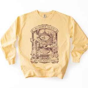 Peut inclure: Un sweat-shirt à col rond jaune pâle avec une impression graphique marron foncé représentant un alchimiste à un bureau avec une loupe, une balance et une plume. Le texte indique ALCHIMIA SILICII et WE TAUGHT THE ROCK TO THINK en bannière.