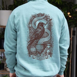 May include: A light blue hoodie featuring a dark brown graphic on the back. The design depicts a raven perched on a cauldron, a book labeled CODEX BINARII, an hourglass, gears, and a banner with the text OPUS MAGNUM, all surrounded by foliage.