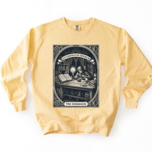 May include: A pale yellow crewneck sweatshirt featuring a black and white graphic print of a hooded figure at a desk with a candle, books, and a magnifying glass. The text reads EXTERMINATOR ERRORUM and THE DEBUGGER in a decorative frame.