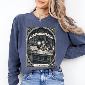Pode incluir: Uma camisola azul de manga comprida com uma impressão gráfica retangular de uma figura encapuzada numa secretária com uma vela, livros e um inseto grande. O texto diz EXTERMINATOR ERRORUM e THE DEBUGGER numa moldura decorativa.
