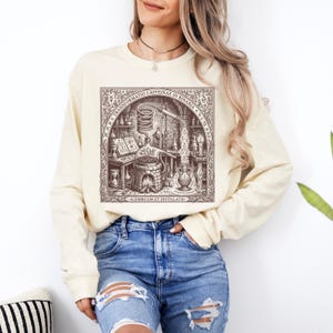 May include: A cream-coloured long-sleeve sweatshirt featuring a brown graphic print of an alchemical laboratory scene. The illustration depicts a distillation apparatus, books, and candles, with the text "TRANSFORMATIO CAFFEINAE IN BINARIA" and "ALEMBICUM ET DISTILLATIO" printed within a decorative border.