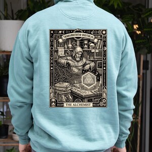Op de afbeelding: Een lichtblauwe hoodie met een gedetailleerde zwart-wit illustratie op de achterkant. De afbeelding toont een figuur met een kap die aan een bureau werkt met wetenschappelijke apparatuur, een geometrische vorm en de tekst Transmutatio Digitalis.