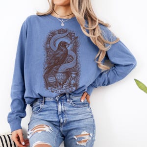 May include: A blue long-sleeve crewneck sweatshirt featuring a dark brown graphic print of a raven perched on a cauldron, surrounded by gears, an hourglass, and a book labeled Codex Binarii, with the text Opus Magnum printed at the bottom.