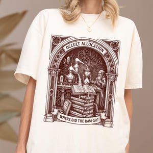 May include: A cream-colored t-shirt featuring a dark brown graphic print of an alchemist in a gothic archway. The design includes laboratory glassware, an hourglass, a stack of books labeled with binary code, and the text Occult Allocation and Where Did The Ram Go?