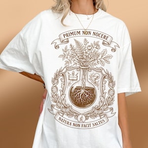 May include: A white t-shirt featuring a brown graphic print of a laboratory flask containing plant roots and flowers. The design includes the Latin text PRIMUM NON NOCERE and NATURA NON FACIT SALTUS, along with scientific symbols like a microscope.